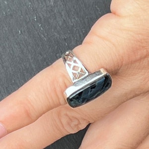 Op de afbeelding: Een zilveren ring met een zwarte steen in een rechthoekige vorm. De band van de ring is ingewikkeld ontworpen met een traliepatroon.