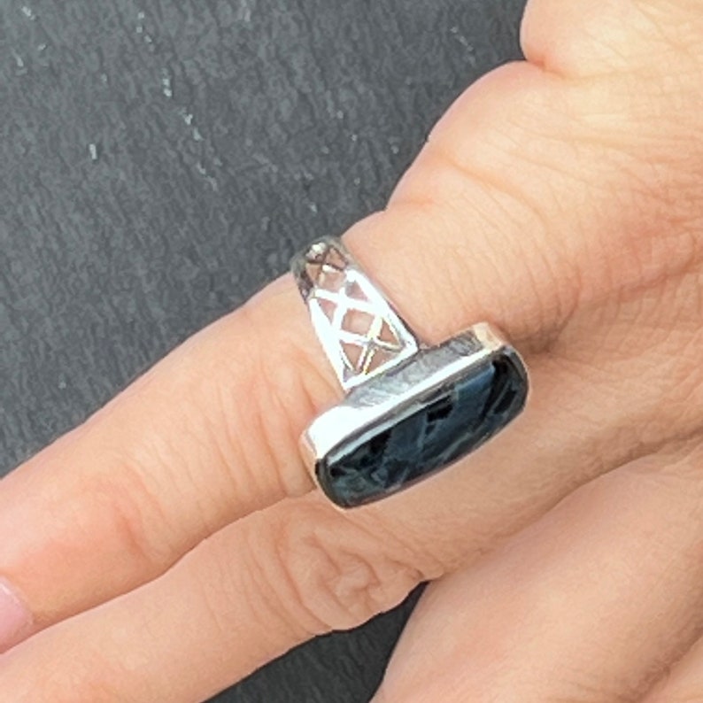 Op de afbeelding: Een zilveren ring met een zwarte steen in een rechthoekige vorm. De band van de ring is ingewikkeld ontworpen met een traliepatroon.