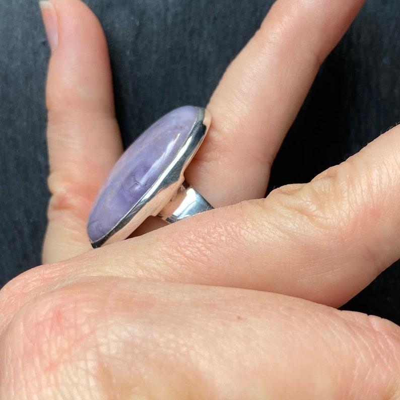 May include: A silver ring with a large, oval-shaped, lavender gemstone set in a bezel setting.