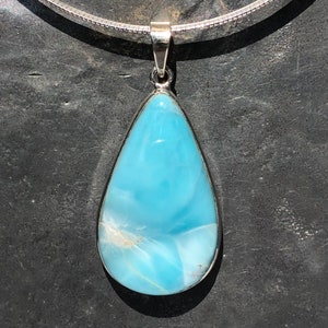 Puede incluir: Un collar con un colgante en forma de lágrima adornado con una piedra preciosa azul claro. El colgante está hecho de plata y tiene un acabado liso y pulido.