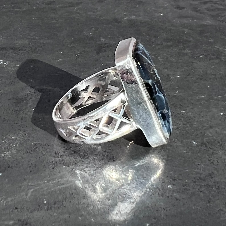 Op de afbeelding: Een zilveren ring met een zwarte en witte steen in een vierkante vorm. De band van de ring is ingewikkeld ontworpen met een traliepatroon.