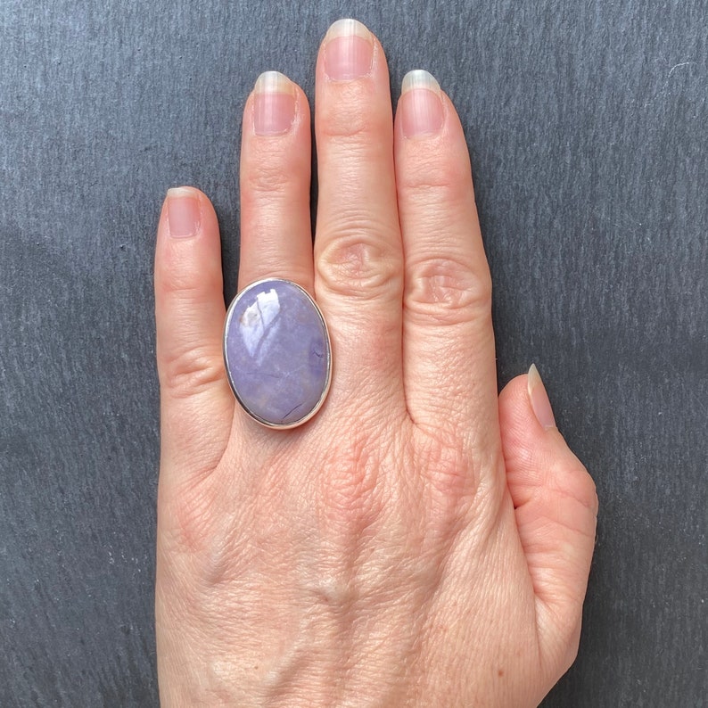 May include: A silver ring with a large oval purple gemstone. The ring is on a person's finger.