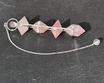 Könnte beinhalten: Ein rosa Rosenquarz-Pendel mit einer silbernen Kette und einem klaren Quarzkristall am Ende. Das Pendel besteht aus fünf Rosenquarz-Pyramiden, die durch eine silberne Stange verbunden sind.