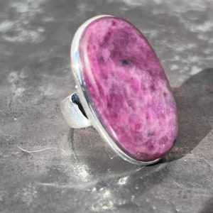 Peut inclure: Une bague imposante avec une grande pierre ovale rose-violette, sertie dans un anneau en argent. La pierre pr&eacute;sente un aspect marbr&eacute; avec des inclusions roses fonc&eacute;es et noires. Le sertissage de la bague est simple et moderne.