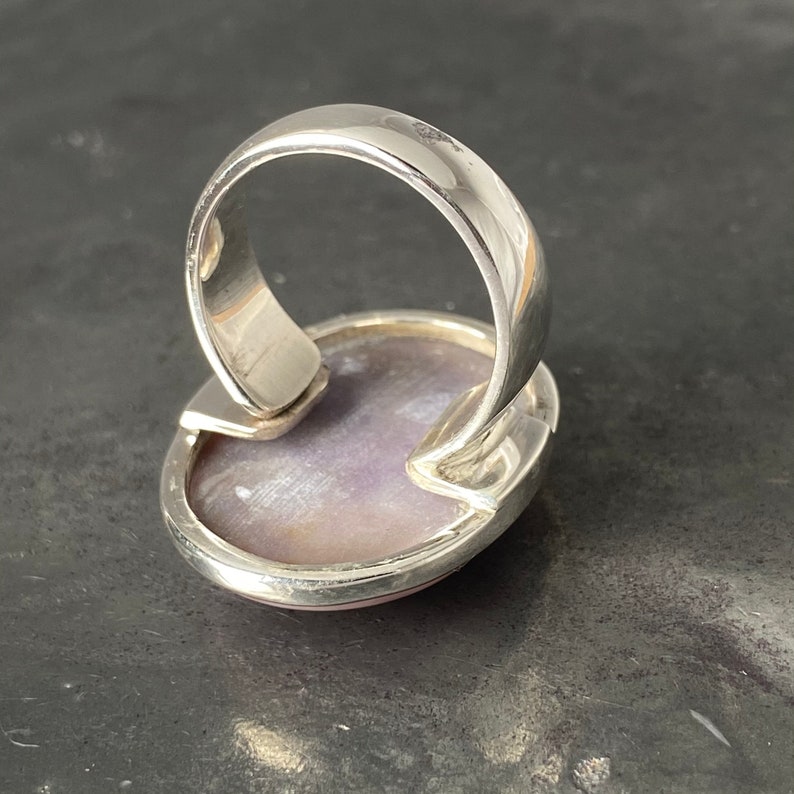 May include: A silver ring with a large oval-shaped purple gemstone set in the center. The ring is made of polished silver and has a simple design.