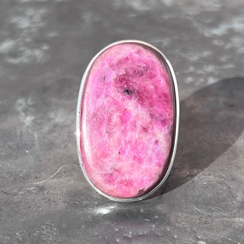 Peut inclure: Bague en pierre rose ovale avec une lunette argent&eacute;e. La pierre a une surface textur&eacute;e avec des inclusions roses fonc&eacute;es et noires. La bague est pos&eacute;e sur une surface grise.