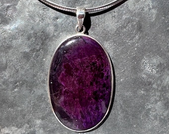 May include: Oval-shaped pendant with a deep purple stone set in a silver-colored metal frame. The pendant hangs from a silver-colored chain. The stone has a marbled appearance with dark and light purple hues.
