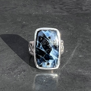 Op de afbeelding: Een zilveren ring met een blauwe en zwarte tijgeroog edelsteen. De ring heeft een rechthoekige zetting en een gedetailleerde band.
