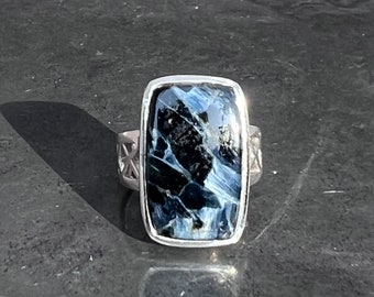 May include: A silver ring with a blue and black tiger's eye gemstone. The ring has a rectangular setting and a detailed band.