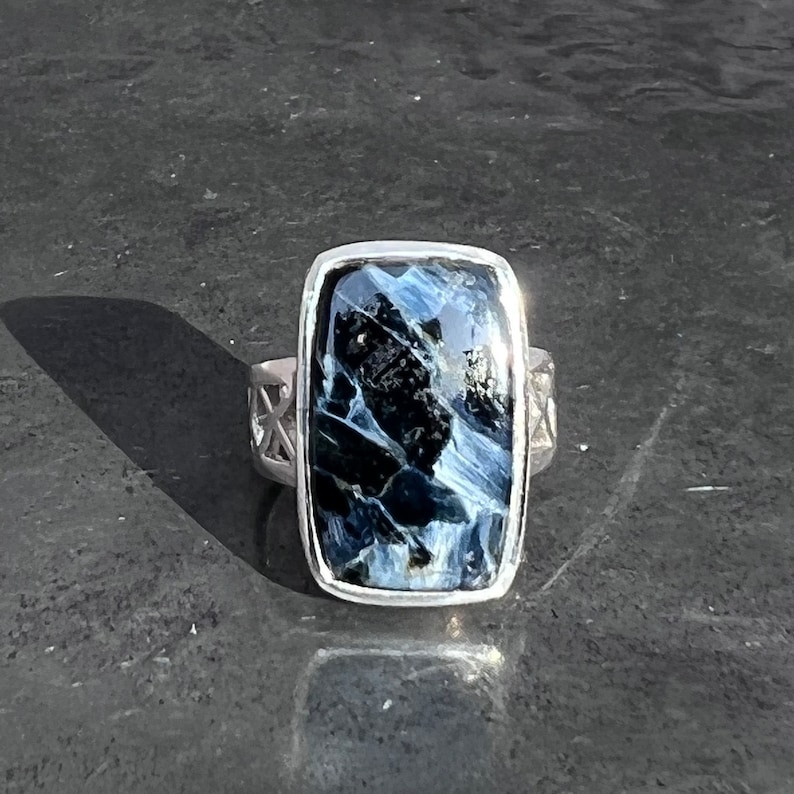 Op de afbeelding: Een zilveren ring met een blauwe en zwarte tijgeroog edelsteen. De ring heeft een rechthoekige zetting en een gedetailleerde band.