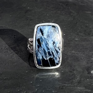 Op de afbeelding: Een zilveren ring met een rechthoekige blauwe en zwarte edelsteen. De edelsteen heeft een uniek patroon van witte en blauwe strepen.
