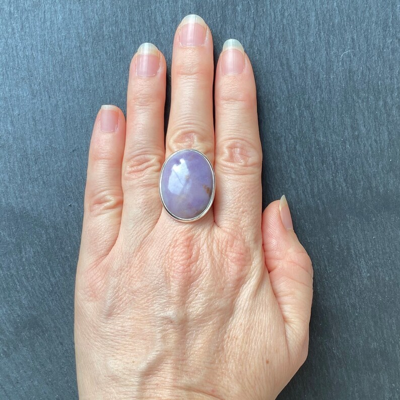 May include: A silver ring with a large, oval-shaped, lavender gemstone. The gemstone has a light purple hue with white and brown inclusions.