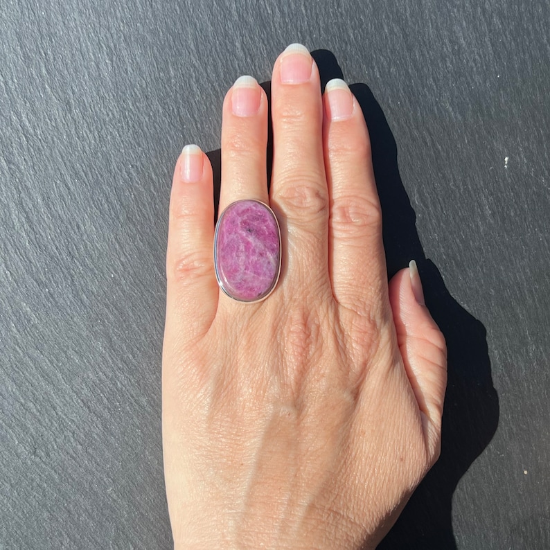 Peut inclure: Une bague ovale avec une pierre pr&eacute;cieuse rose vif sertie dans un anneau argent&eacute;. La pierre a une surface textur&eacute;e avec des inclusions roses plus fonc&eacute;es. La bague est pr&eacute;sent&eacute;e sur une main sur fond sombre.