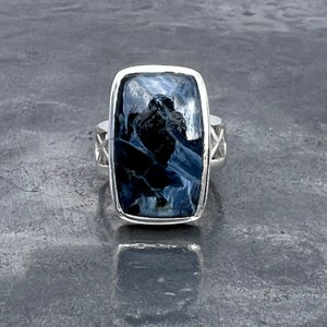 Op de afbeelding: Een zilveren ring met een rechthoekige blauwe en zwarte edelsteen. De edelsteen heeft een draaiend patroon.