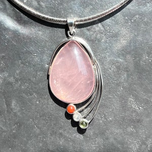 Könnte beinhalten: Eine silberne Halskette mit einem Anhänger aus rosa Rosenquarz. Der Anhänger ist tropfenförmig und hat eine silberne Fassung. Die Halskette hat außerdem drei kleinere Edelsteine in Rot, Weiß und Grün.
