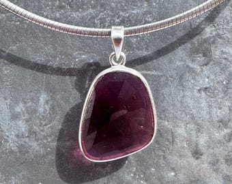 Op de afbeelding: Een zilveren hangerketting met een gefacetteerde, dieppaarse edelsteen. De hanger heeft een onregelmatige vorm en is gezet in een zilveren bezel. De ketting is een dunne, flexibele zilveren slangenketting.