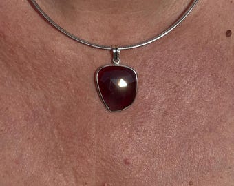 Op de afbeelding: Een zilveren ketting met een hanger met een gefacetteerde, dieprode edelsteen. De hanger is gezet in een zilveren rand en hangt aan een dunne, zilveren ketting. De edelsteen heeft een driehoekige vorm.