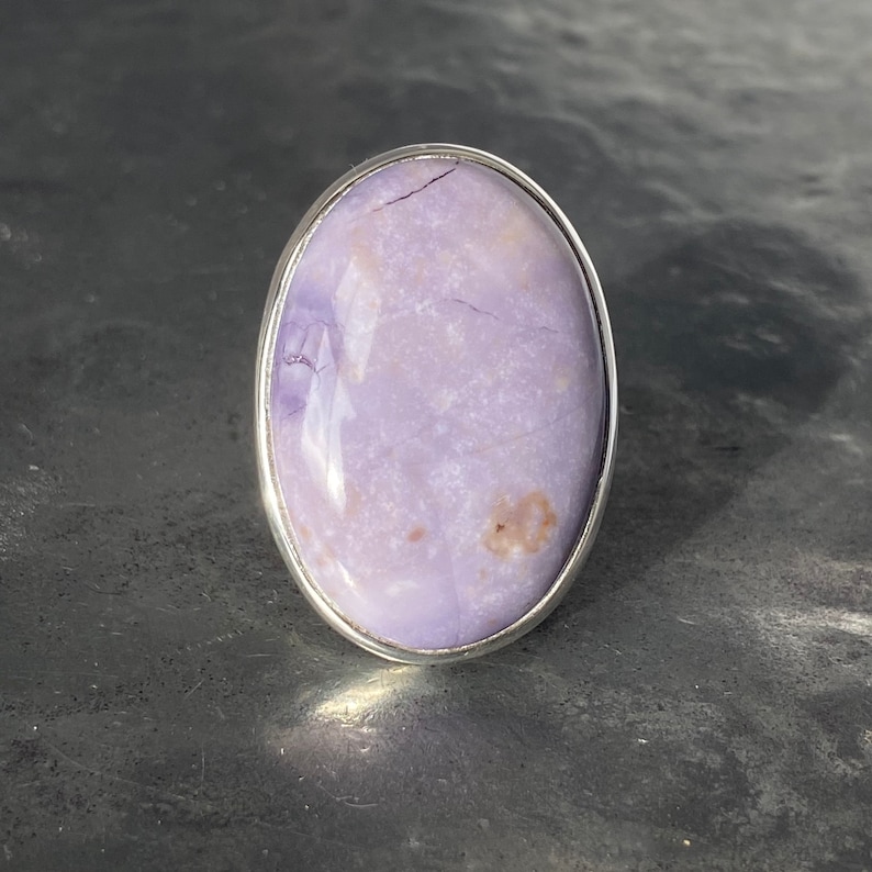May include: A silver ring with a large oval-shaped purple gemstone. The gemstone has a marbled pattern and a light purple hue.