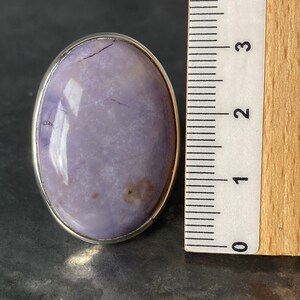 May include: A silver ring with a large oval-shaped purple gemstone. The stone has a cloudy, marbled appearance with a few darker brown spots. The ring is shown next to a ruler, measuring approximately 1 inch in diameter.