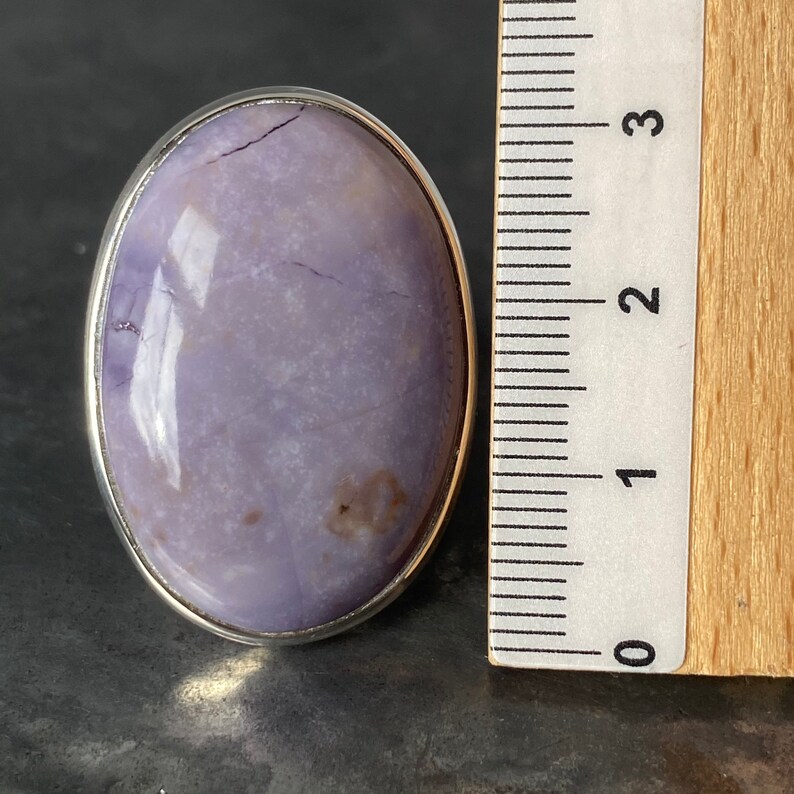 May include: A silver ring with a large oval-shaped purple gemstone. The stone has a cloudy, marbled appearance with a few darker brown spots. The ring is shown next to a ruler, measuring approximately 1 inch in diameter.