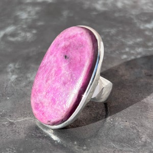 Peut inclure: Une bague ovale avec une grosse pierre rose vif sertie dans un anneau en argent poli. La pierre pr&eacute;sente une surface textur&eacute;e avec de subtiles variations de couleur. Le design de la bague est simple mais saisissant, adapt&eacute; au quotidien ou aux occasions sp&eacute;ciales.