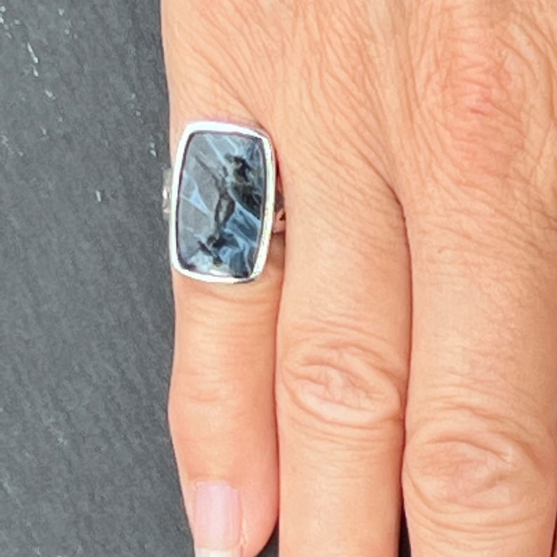 Op de afbeelding: Een zilveren ring met een rechthoekige blauwe en grijze steen in het midden.