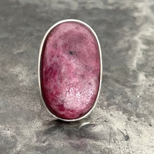 Peut inclure: Bague ovale avec une grosse pierre rose polie, sertie dans un sertissage argent&eacute;. La pierre pr&eacute;sente un motif mouchet&eacute; avec des nuances de rose. La bague est pr&eacute;sent&eacute;e sur un fond gris textur&eacute;.