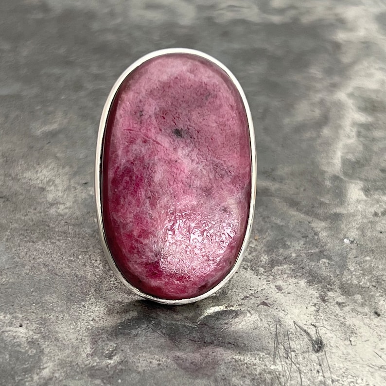 Peut inclure: Bague ovale avec une grosse pierre rose polie, sertie dans un sertissage argent&eacute;. La pierre pr&eacute;sente un motif mouchet&eacute; avec des nuances de rose. La bague est pr&eacute;sent&eacute;e sur un fond gris textur&eacute;.