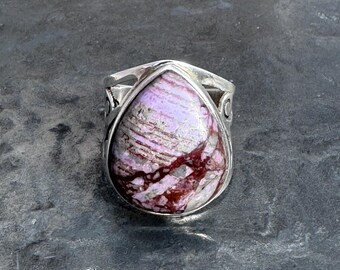 May include: A silver ring with a large, teardrop-shaped stone. The stone is a mix of lavender, burgundy, and white. The ring's band has decorative cutouts on the sides.