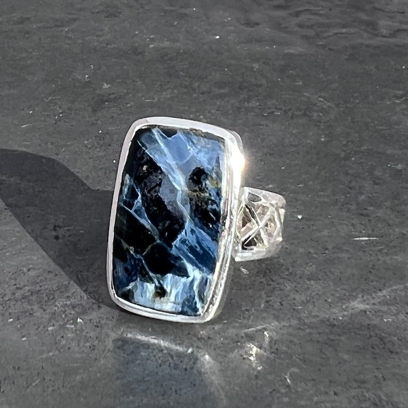 Op de afbeelding: Een zilveren ring met een rechthoekige blauwe en zwarte tijgeroog-edelsteen. De ring heeft een gedetailleerde zilveren band.