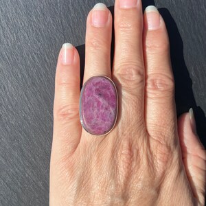 Peut inclure: Une bague ovale avec une pierre pr&eacute;cieuse rose vif sertie dans un anneau argent&eacute;. La pierre a une surface textur&eacute;e avec des inclusions roses fonc&eacute;es et noires. La bague est pr&eacute;sent&eacute;e sur une main, mettant en valeur sa taille et sa couleur.