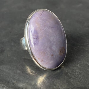 May include: A silver ring with a large oval-shaped purple gemstone. The gemstone has a cloudy, translucent appearance with white and brown inclusions.