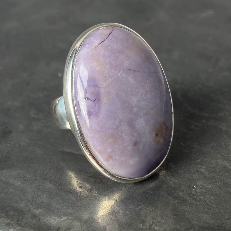 May include: A silver ring with a large oval-shaped purple gemstone. The gemstone has a cloudy, translucent appearance with white and brown inclusions.