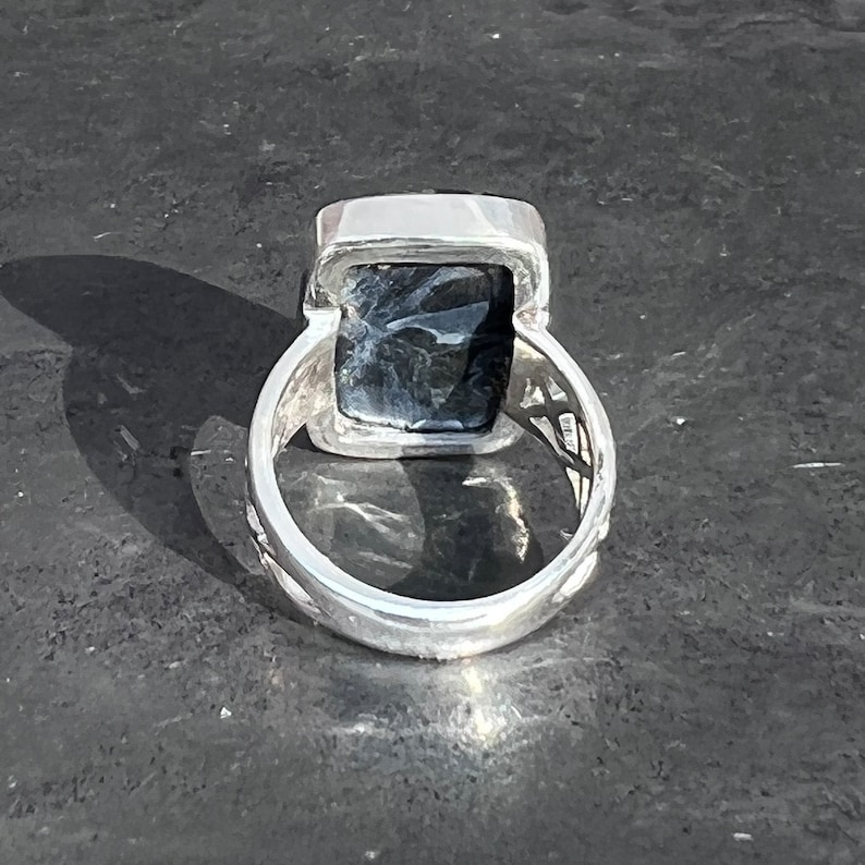 Op de afbeelding: Een zilveren ring met een donkergrijze rechthoekige steen in een bezel-setting. De band van de ring is getextureerd en heeft een decoratief ontwerp.