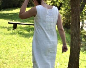 May include: A woman wearing a white maxi dress walks through a grassy area with a tree in the background.