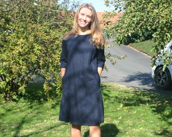 May include: A woman wearing a dark blue knee-length dress with long sleeves and pockets. She is standing on a grassy lawn with trees in the background.