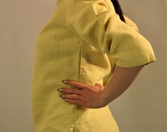 May include: A pale yellow linen shirt with a slightly loose fit and three-quarter sleeves. The shirt has a simple, rounded neckline and a side seam detail.