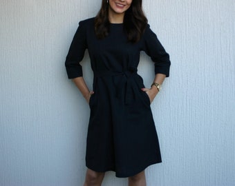 May include: A woman wearing a black dress with a tie belt and pockets. She is standing in front of a white wall.