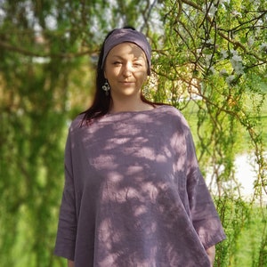 May include: A woman wearing a loose-fitting, mauve linen top with a headband. She is standing in front of a green leafy background.