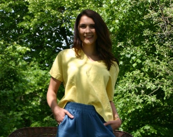 May include: A woman wearing a yellow linen top and blue denim pants. She is standing in front of a green leafy background.