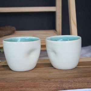 May include: Two white ceramic mugs with a light blue interior. The mugs have a unique, textured exterior.