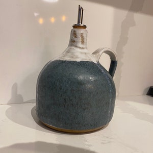 May include: A ceramic oil cruet with a dark blue glazed body and a white upper section. It has a small handle and a metal spout. The cruet is designed for dispensing oil and other liquids.