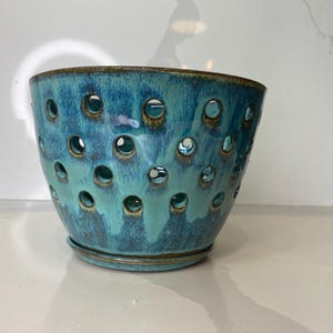 Peut inclure: Un pot de fleur en céramique turquoise percé de nombreux trous circulaires. Le pot a une forme de bol large et ouvert avec un bord légèrement évasé. L'émail a une finition brillante avec des accents bleu foncé et marron. Une soucoupe assortie est attachée au fond.