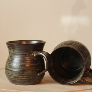 May include: Two brown ceramic mugs with green glaze. The mugs have a rounded body and a handle. The mugs are sitting on a wooden surface.