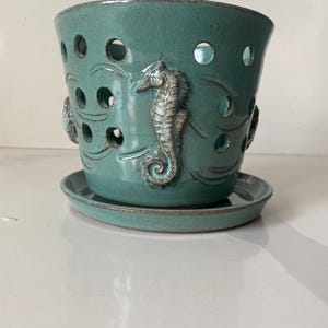 May include: Teal ceramic planter with a matching saucer. The planter features a seahorse and wave design, with circular cutouts. The planter is approximately 6 inches tall and 6 inches wide.