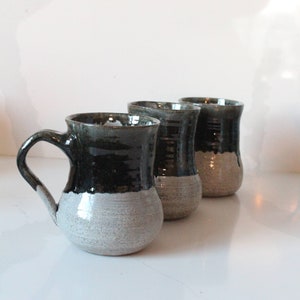 May include: Three ceramic mugs with a black and white glaze. The mugs have a rounded shape and a handle.