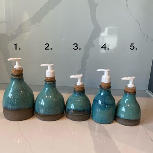 May include: Five ceramic soap dispensers with a teal glaze and brown base. Each bottle has a white pump and a cork stopper. The dispensers are arranged in a row, varying slightly in size and shape.