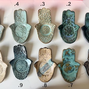 May include: Ten ceramic hamsa-shaped trinket dishes arranged in two rows, numbered 1 through 10. Each dish features unique embossed patterns, including floral, geometric, and textured designs, finished in various glaze colors like blue, green, and tan.