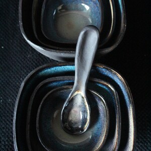 Puede incluir: Un juego de cuatro cuencos de cer&aacute;mica negra y azul con una cuchara de cer&aacute;mica gris oscuro. Los cuencos est&aacute;n anidados uno dentro del otro y tienen un acabado brillante.