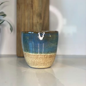 May include: A blue and brown ceramic mug with a speckled glaze. The mug is sitting on a white countertop.